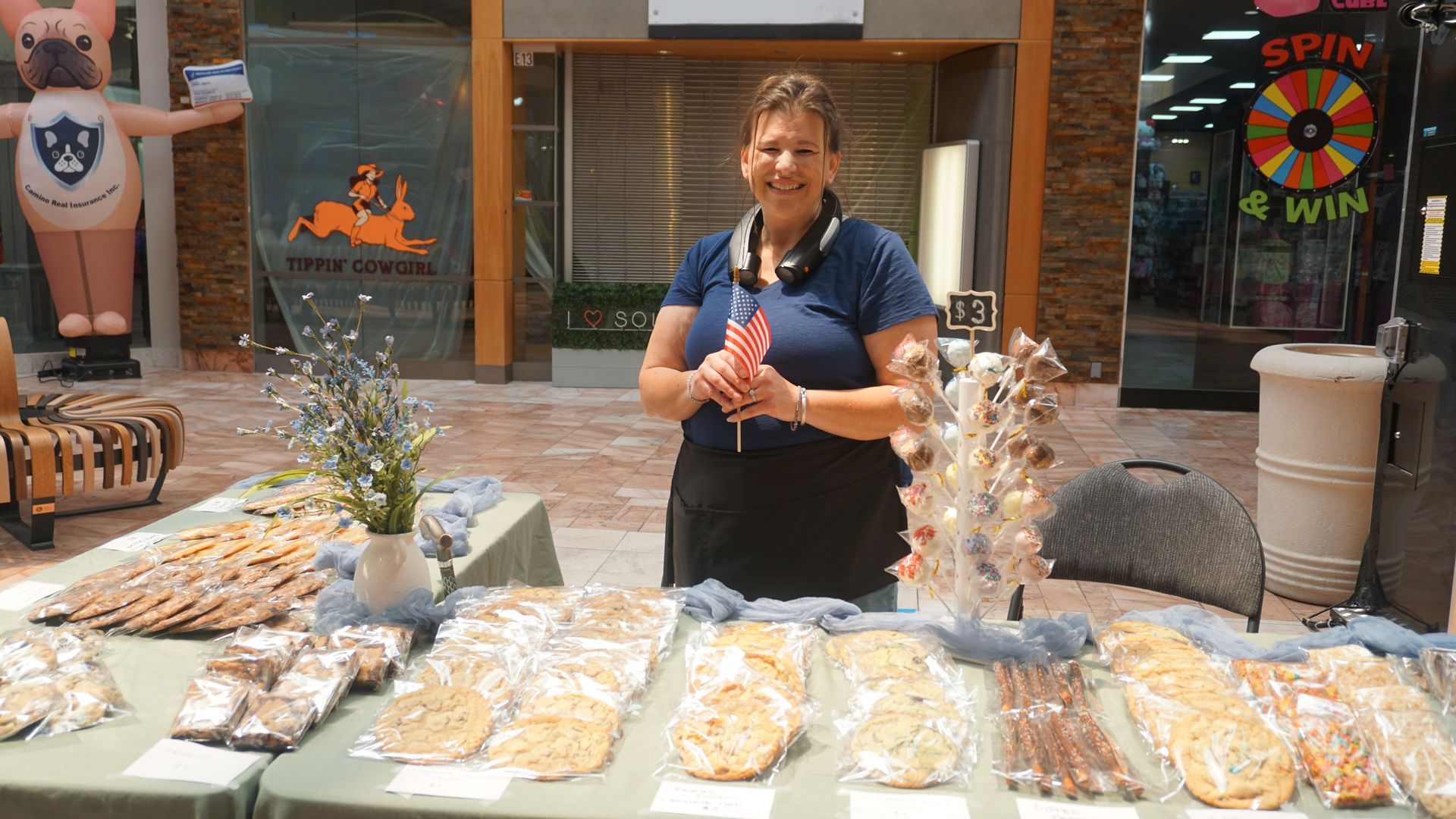 Vendor smiling behind a table of baked goods at an outdoor market stall