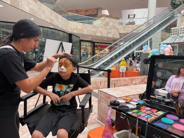 Makeup artist applying face paint to seated person in a mall, with cosmetics displayed nearby.