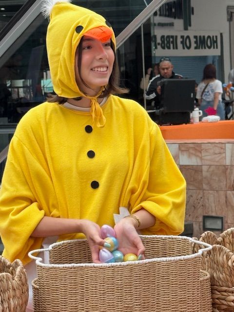 Person in a yellow bird costume holding Easter eggs in a wicker basket outdoors