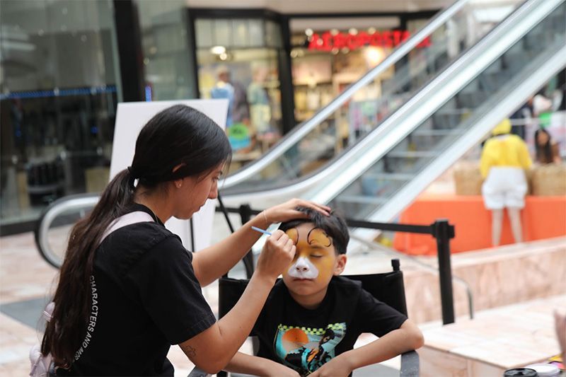 Child getting face paint at an indoor mall table, with escalators in the background.