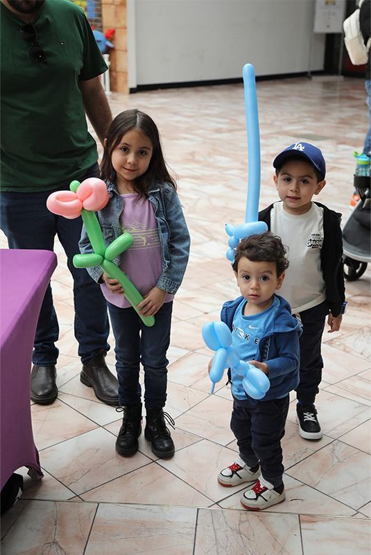 Children holding balloon animals indoors; a girl and two boys stand in a hall with adults nearby.