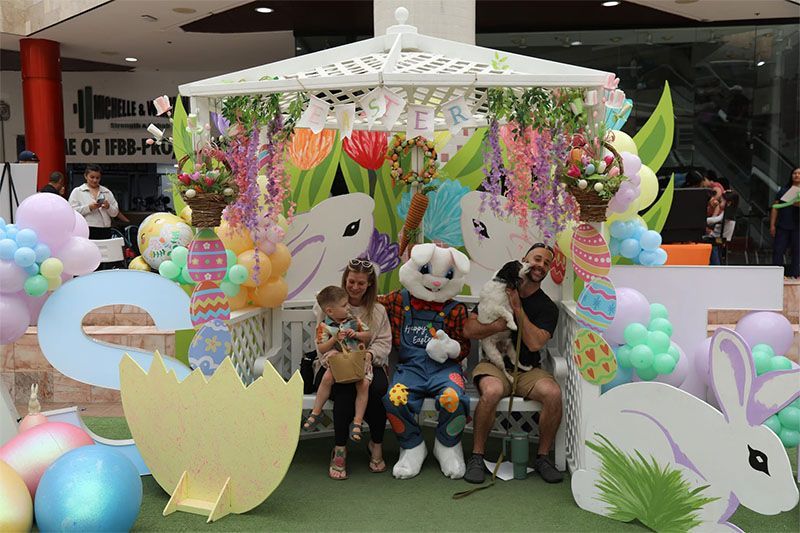 Children posing with two mascot characters at a colorful Easter-themed display with balloons and bunny decorations