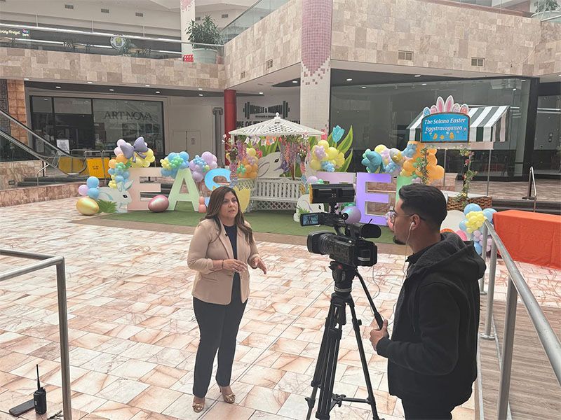 Woman speaking in a mall atrium while a cameraman films her near a colorful balloon display