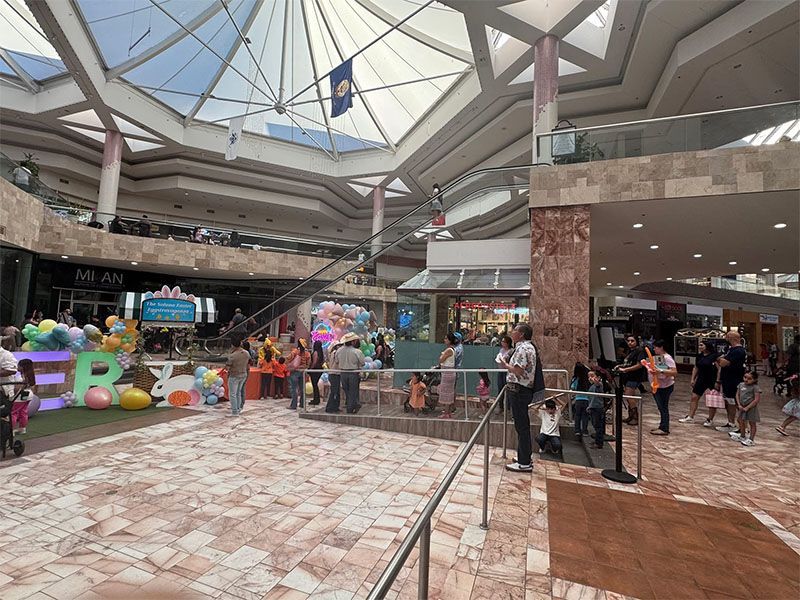 Indoor mall atrium with shoppers, colorful art display, and a glass skylight ceiling