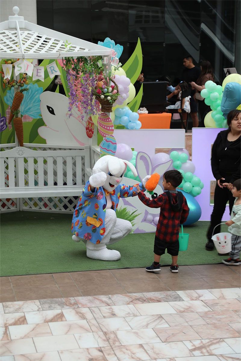 Child in plaid coat high-fives a white costumed mascot beside a decorated floral display.