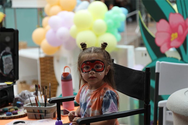 Child wearing red glasses sits at a table with balloons and a flower decoration in the background