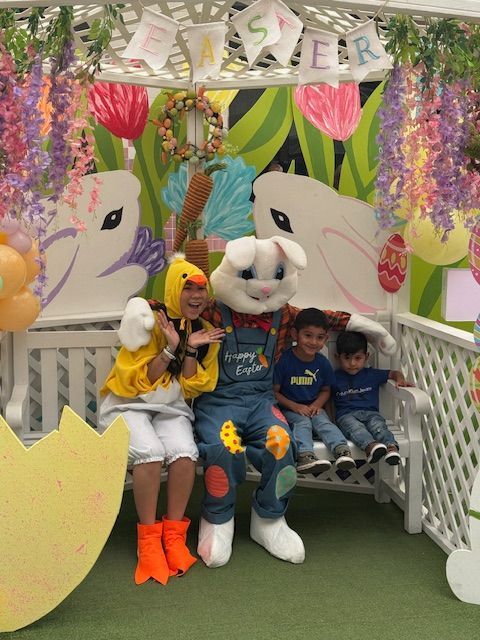 Two children pose with Bugs Bunny and a yellow-costumed character in a colorful floral Easter display.