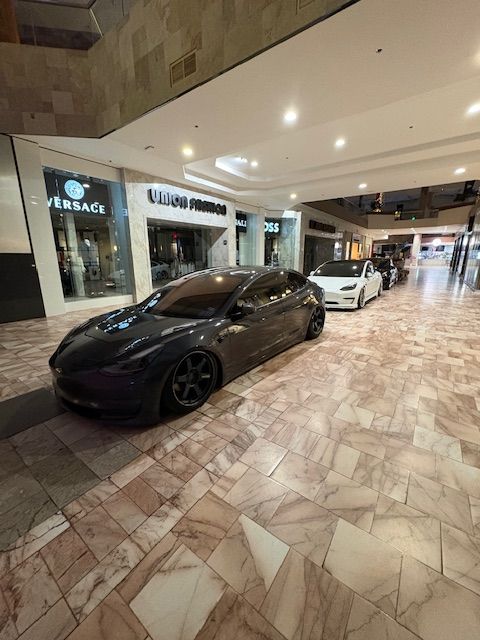 Luxury cars displayed in a mall corridor, including a matte black sports car in the foreground and white car behind.