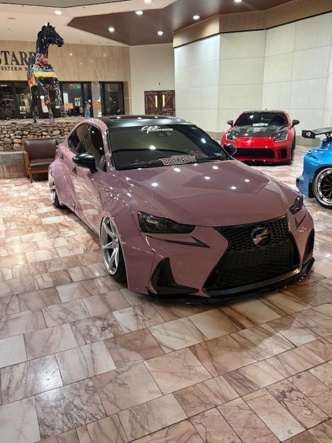Matte pink Lexus sedan displayed indoors on a tiled showroom floor, with red and blue sports cars behind it