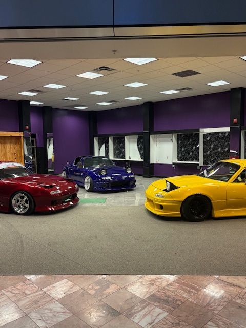 Three sports cars in a showroom: red, blue, and yellow under bright ceiling lights.