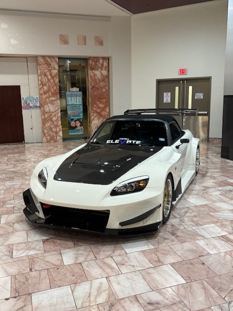 White sports car with black hood displayed indoors on a tiled floor near double doors.