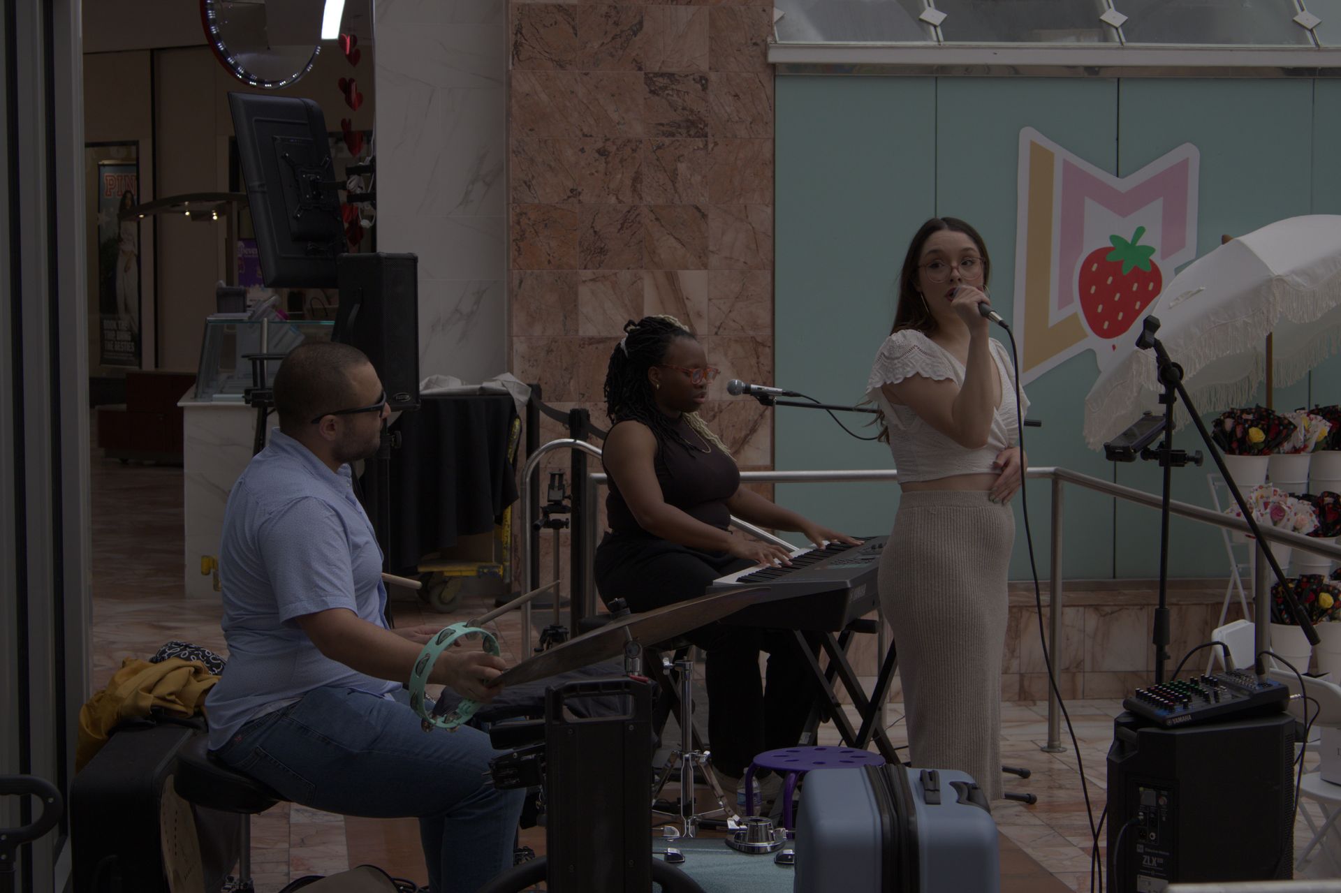 Musicians performing on a small stage, with a singer at a microphone and a drummer and keyboardist beside her.