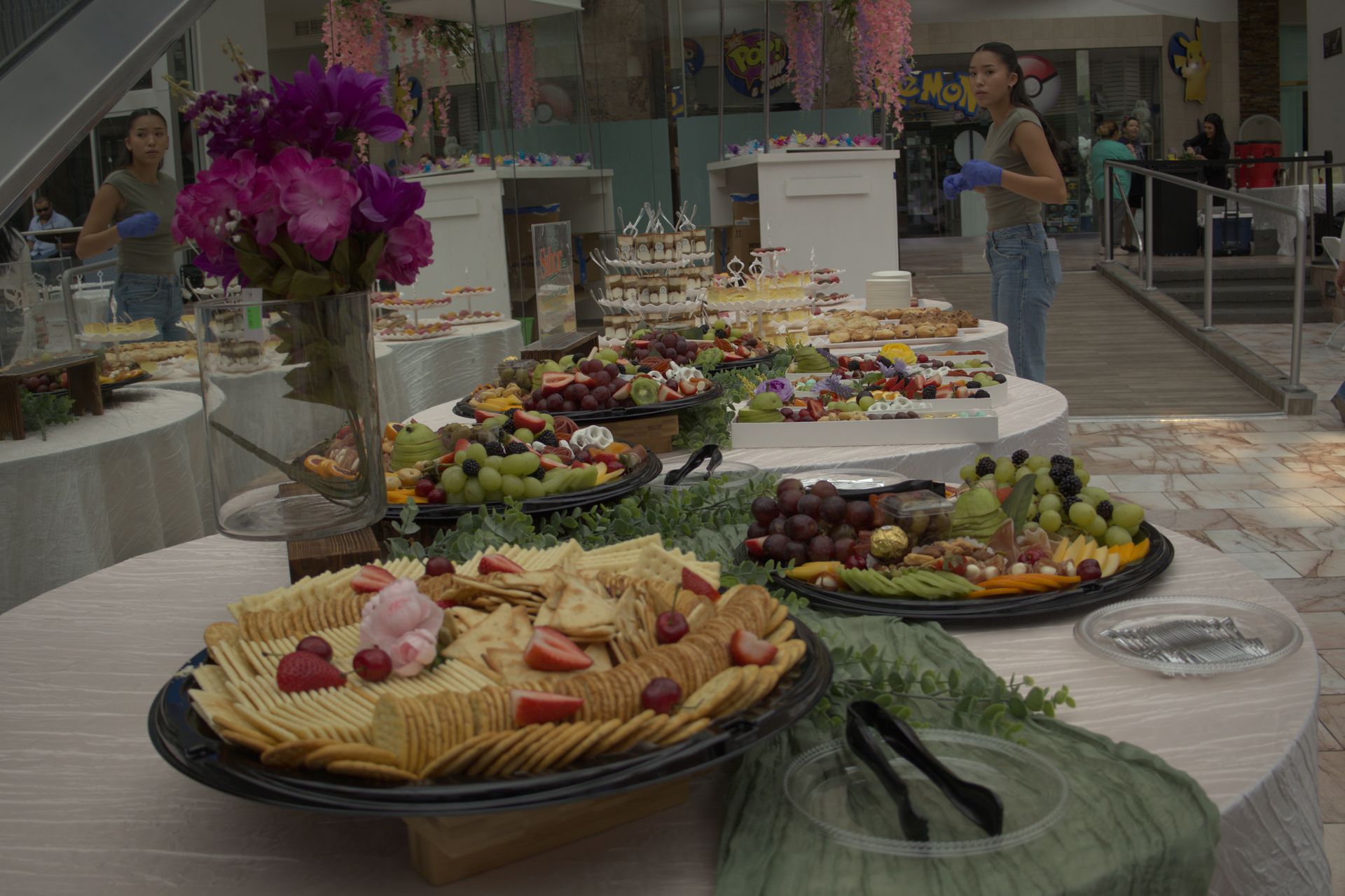 Buffet table with assorted fruit and desserts, including a purple flower centerpiece, in a bright indoor venue
