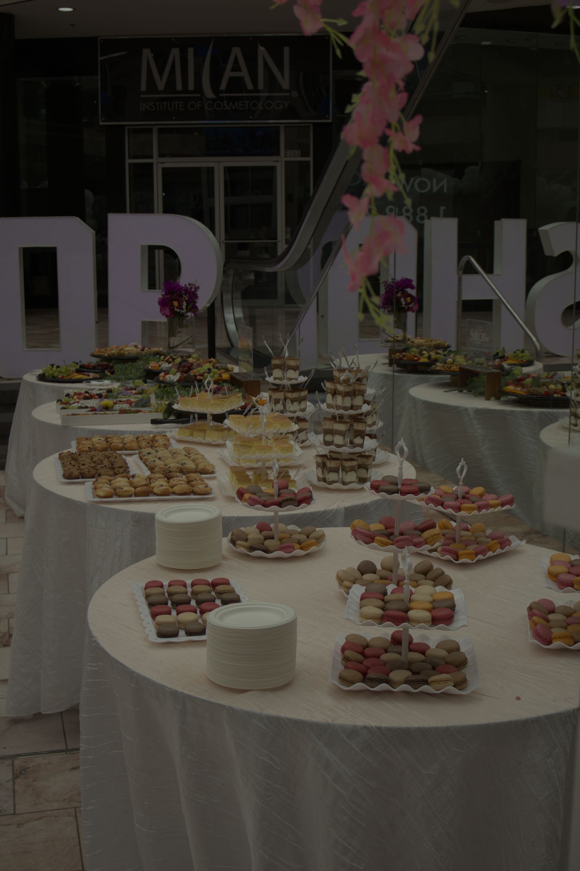 Dessert buffet on round white tables at a reception, with pastries, cakes, and pink floral decor