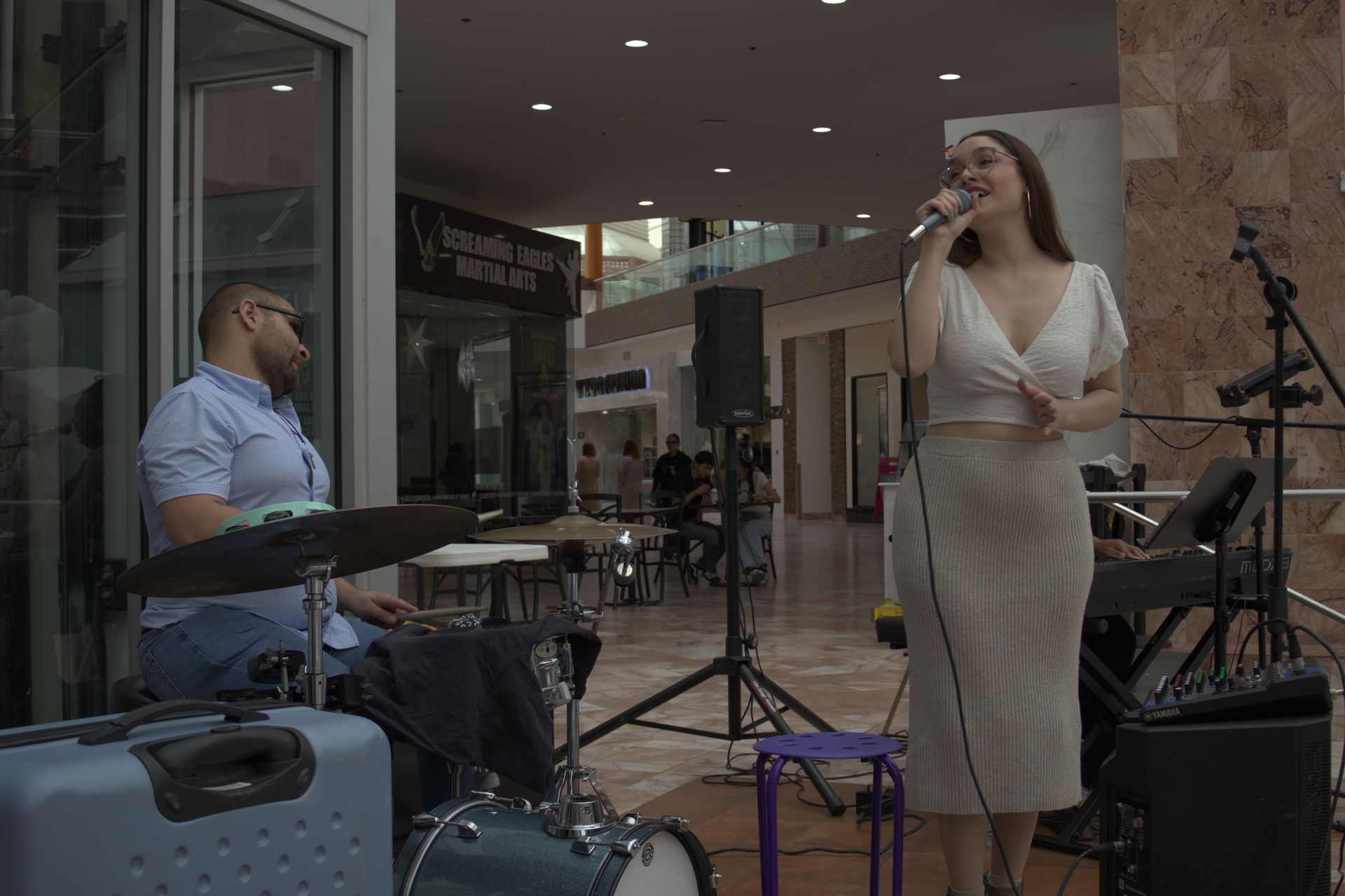 Street musician singing into a mic beside a seated drummer outside a storefront.