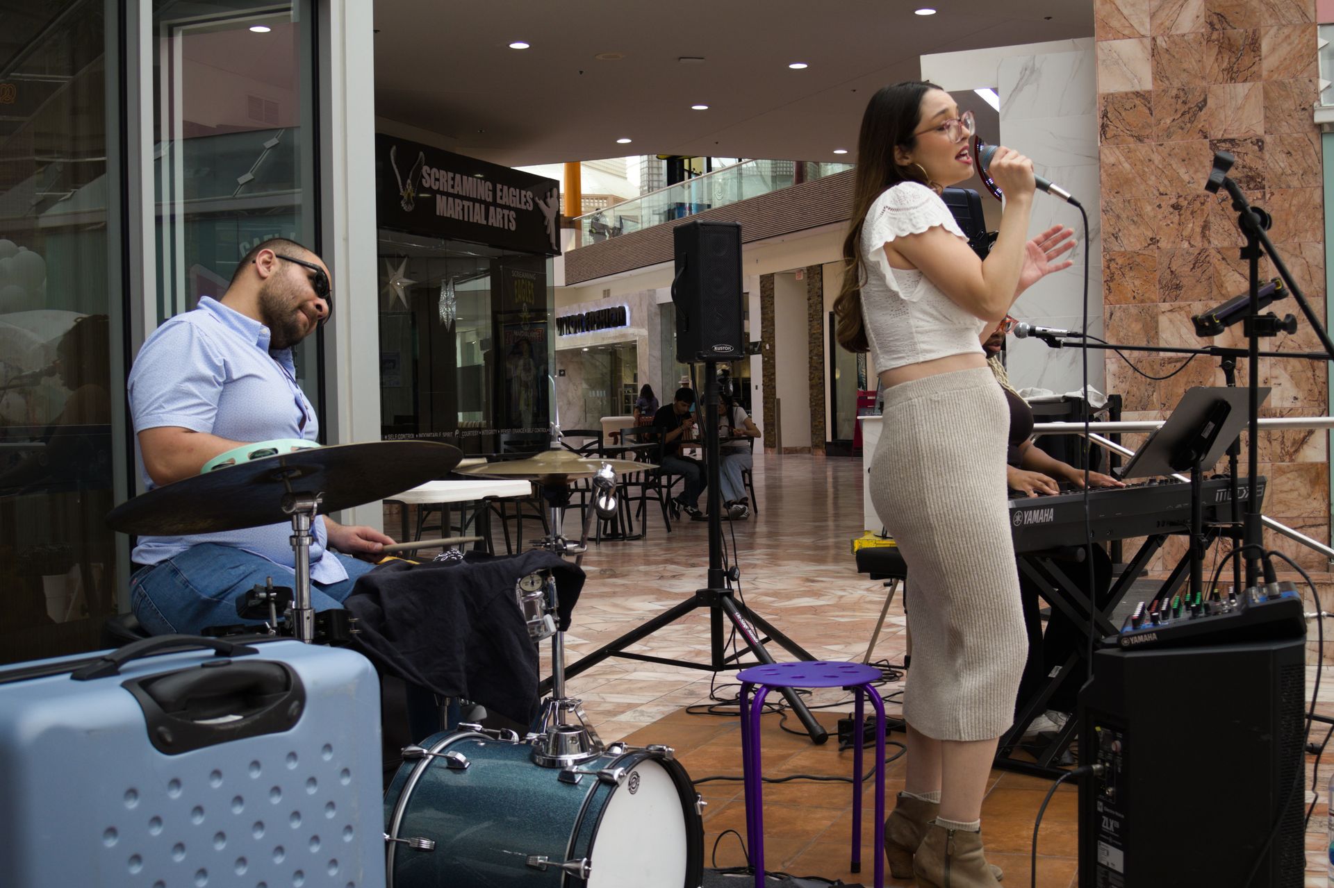 Singer performing beside drummer in an indoor mall, holding a microphone on a small stage.