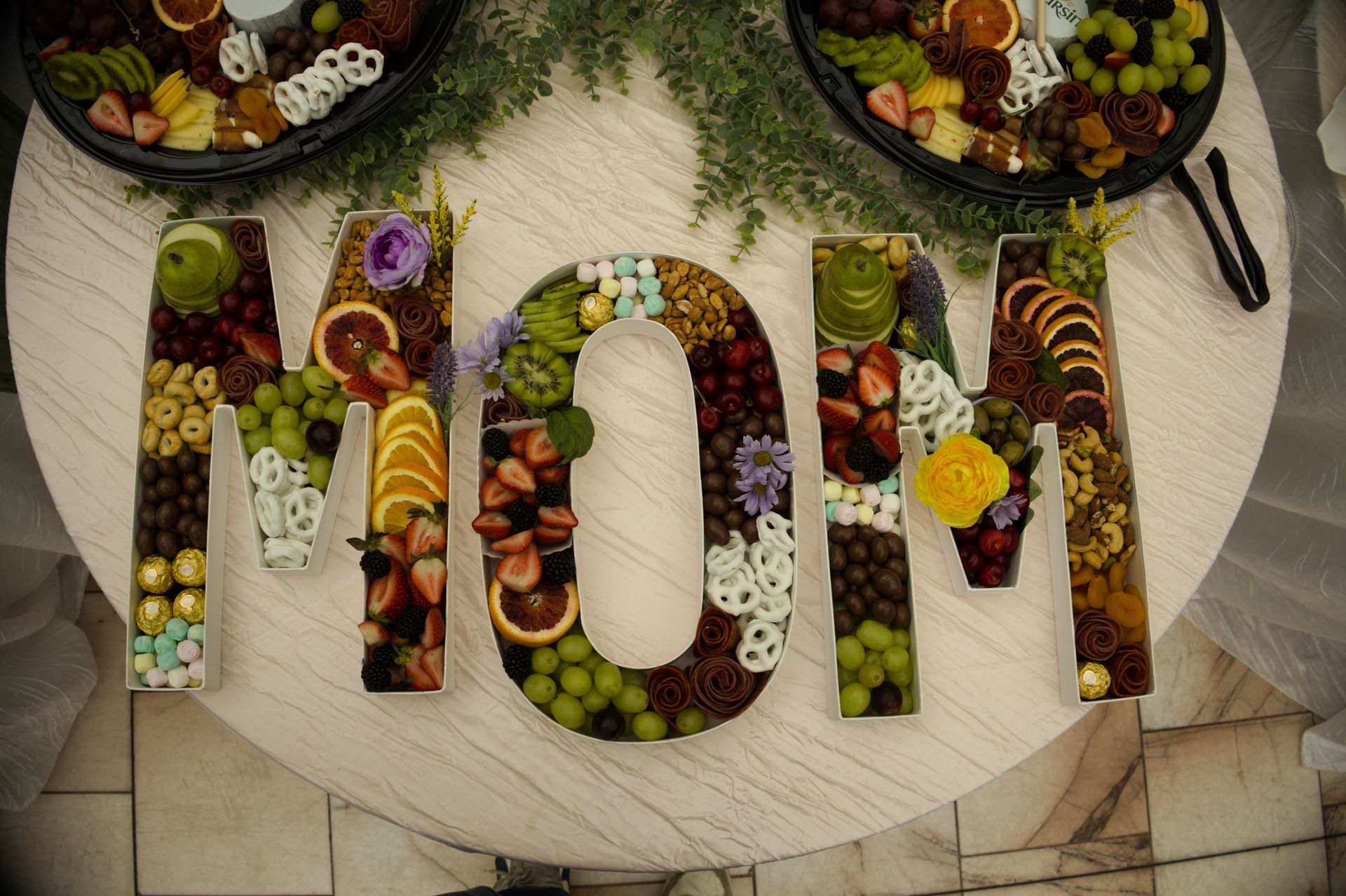 “MOM” spelled with fruit and snacks on a round table, with platters around it.