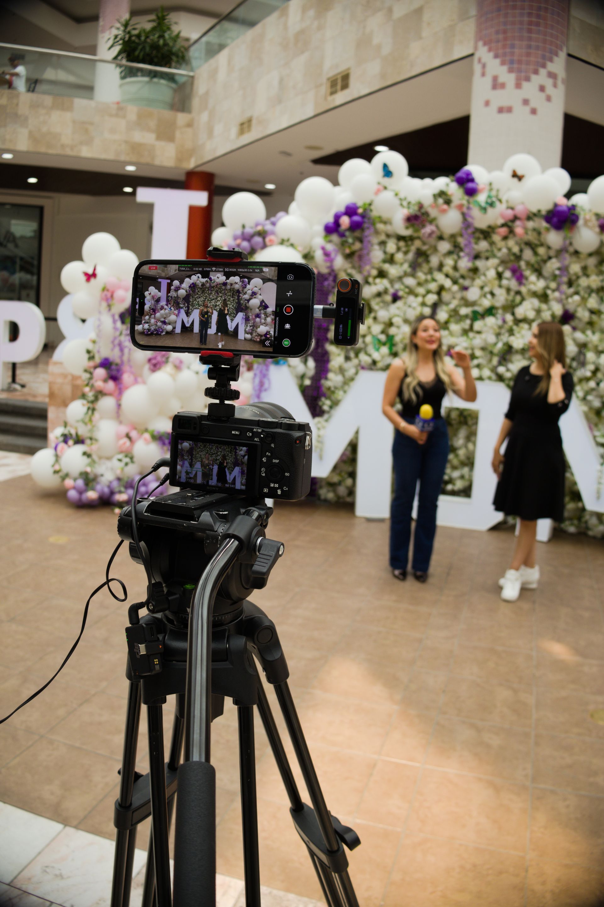 Two women posing in front of a floral balloon backdrop, filmed by a camera on a tripod.
