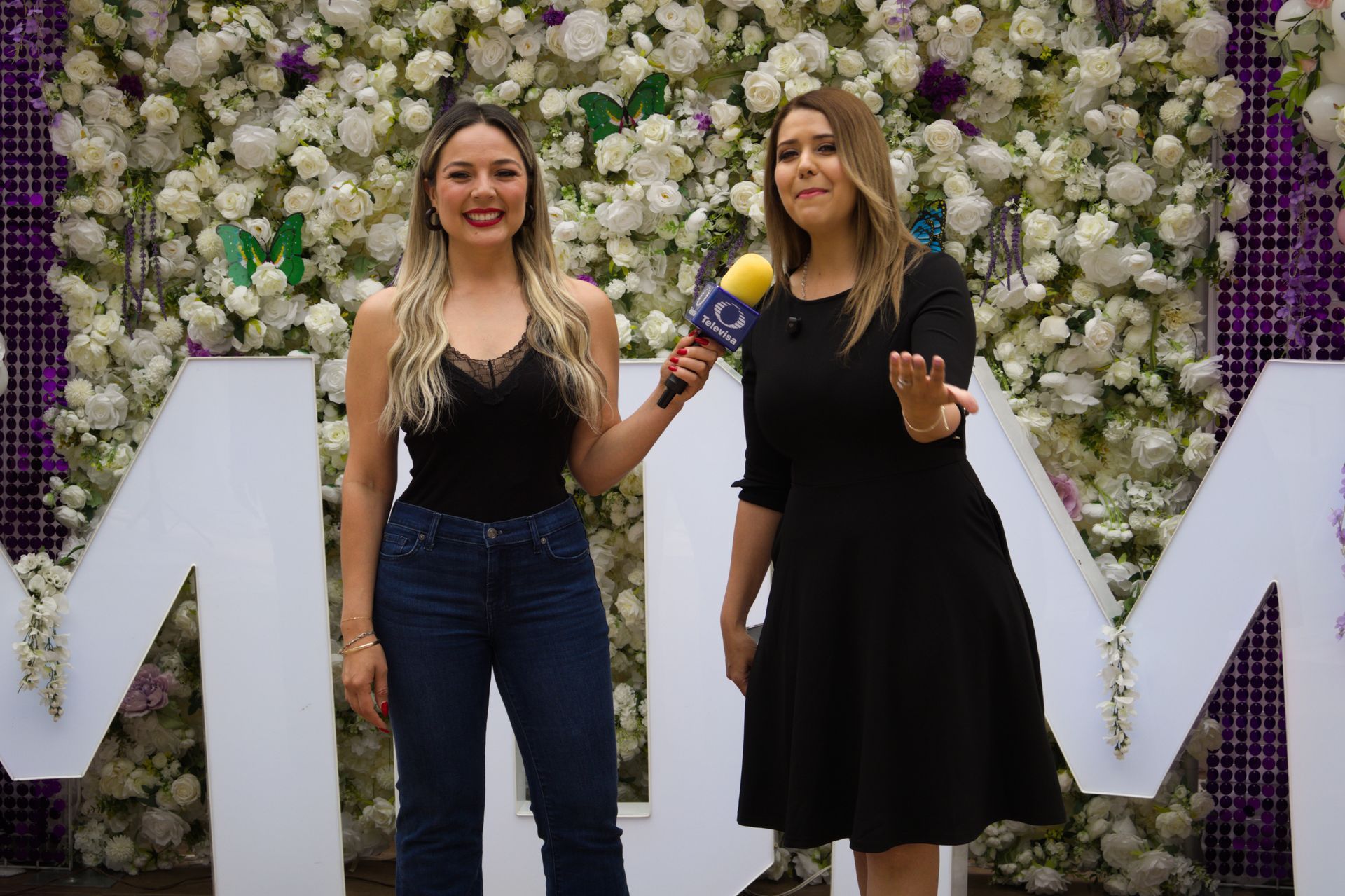 Two women posing in front of a flower wall, one holding a microphone, with large white letters behind them