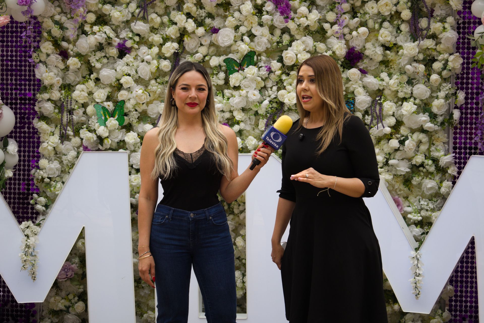 Two women standing before a flower wall, one holding a microphone in a black dress