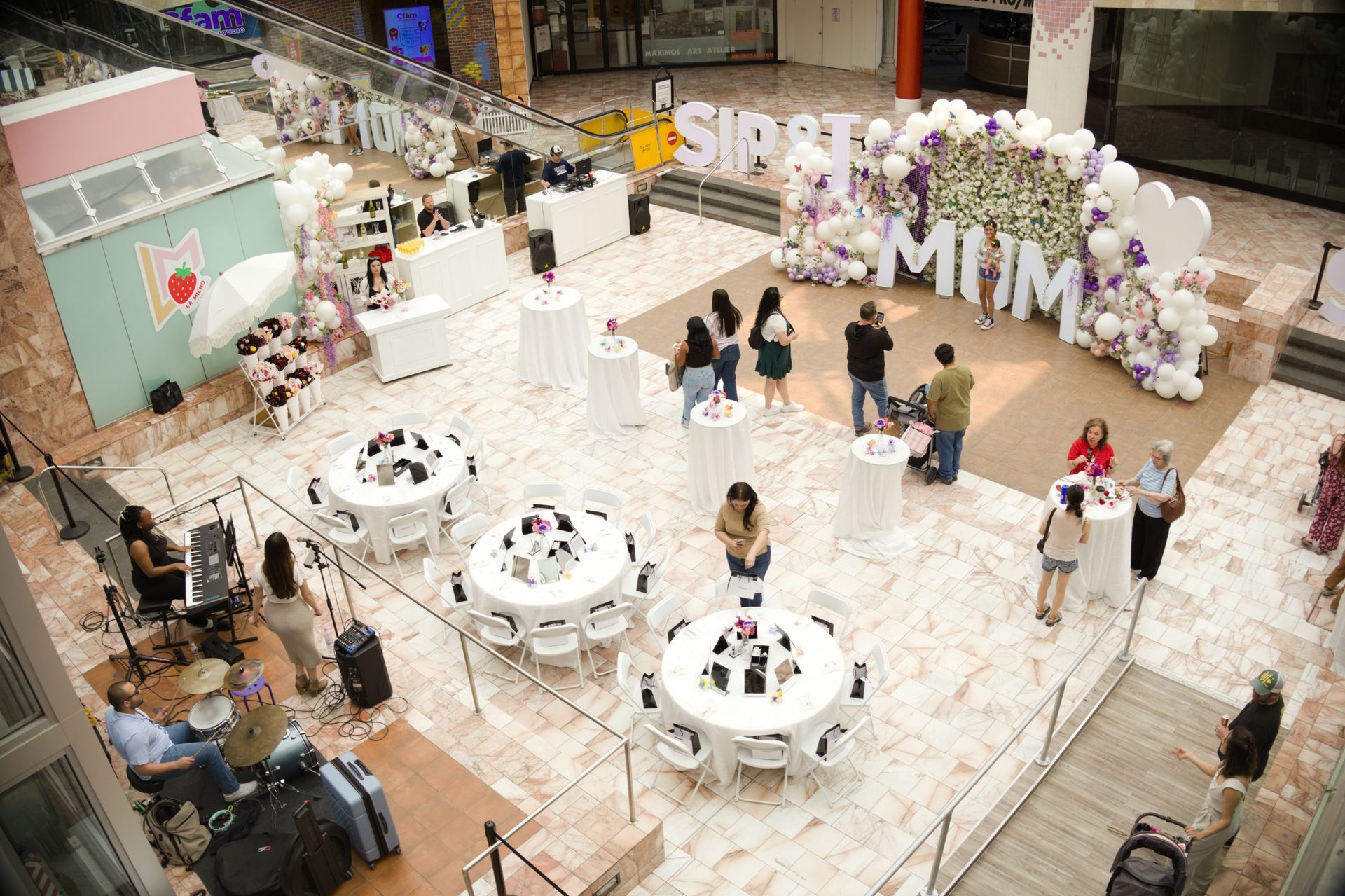 Indoor event space with white tables, guests mingling, and a pink-and-white balloon display reading “MIMI”
