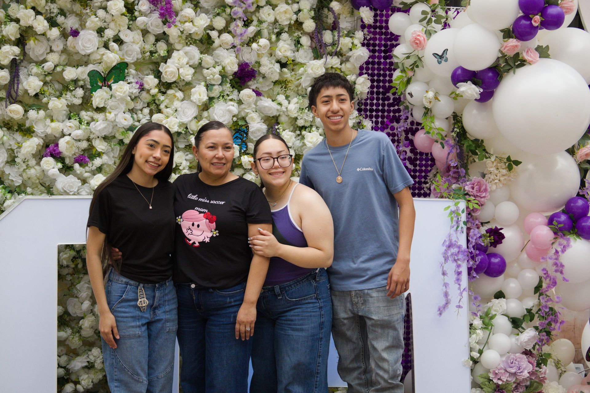 Four people posing in front of a flower wall with purple and white balloon decorations