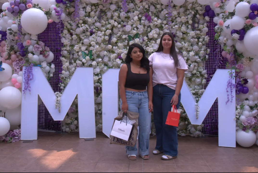 Two women posing in front of giant white M letters and a floral wall with purple balloons