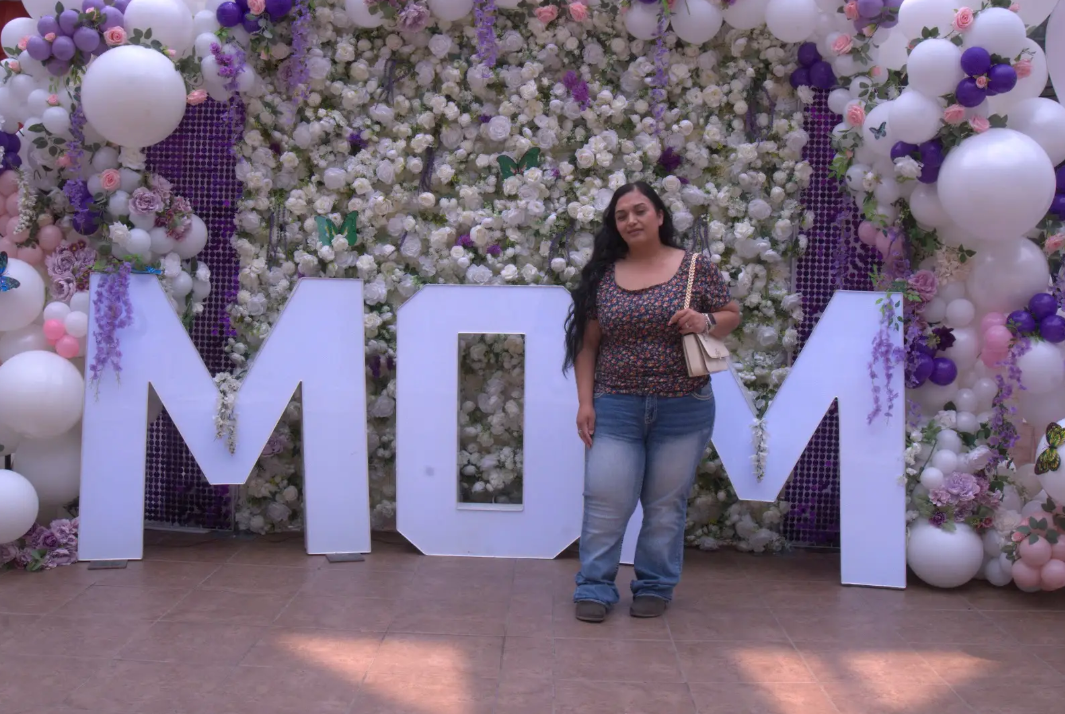 Woman standing in front of large white “MOM” letters and a floral balloon backdrop at an outdoor celebration