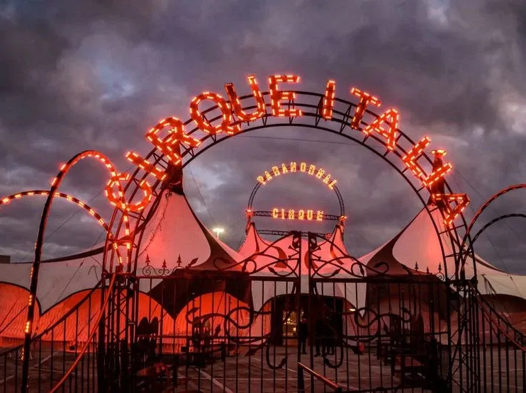 Glowing orange circus tent entrance under a cloudy dusk sky, with arched neon lettering and a fenced gate