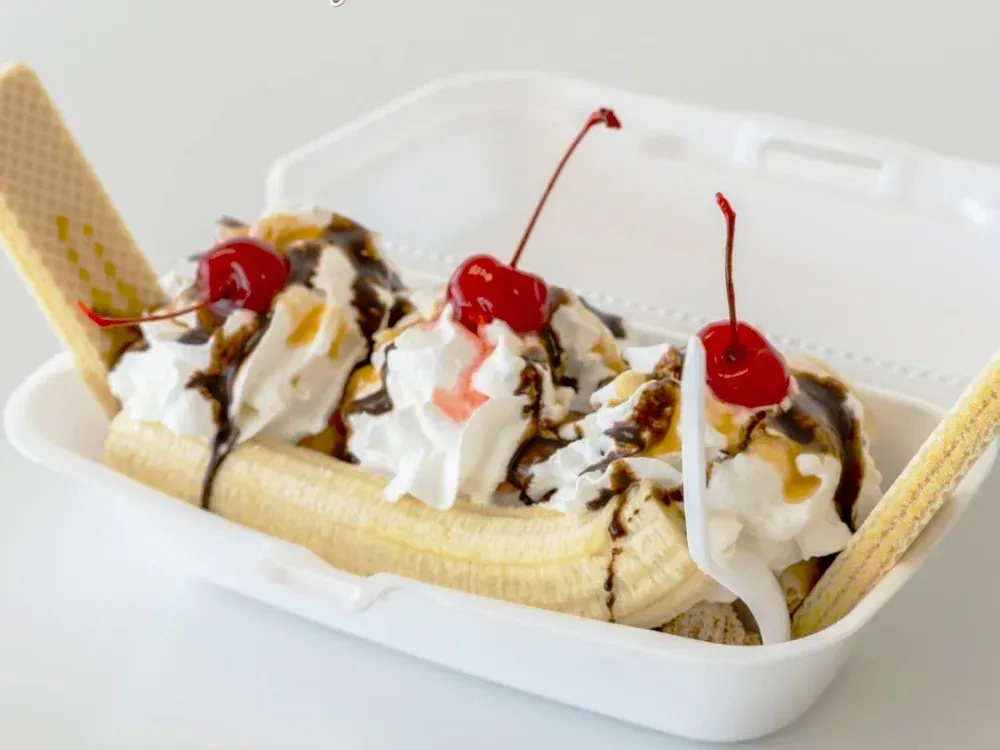 A banana split with whipped cream and cherries in a styrofoam container