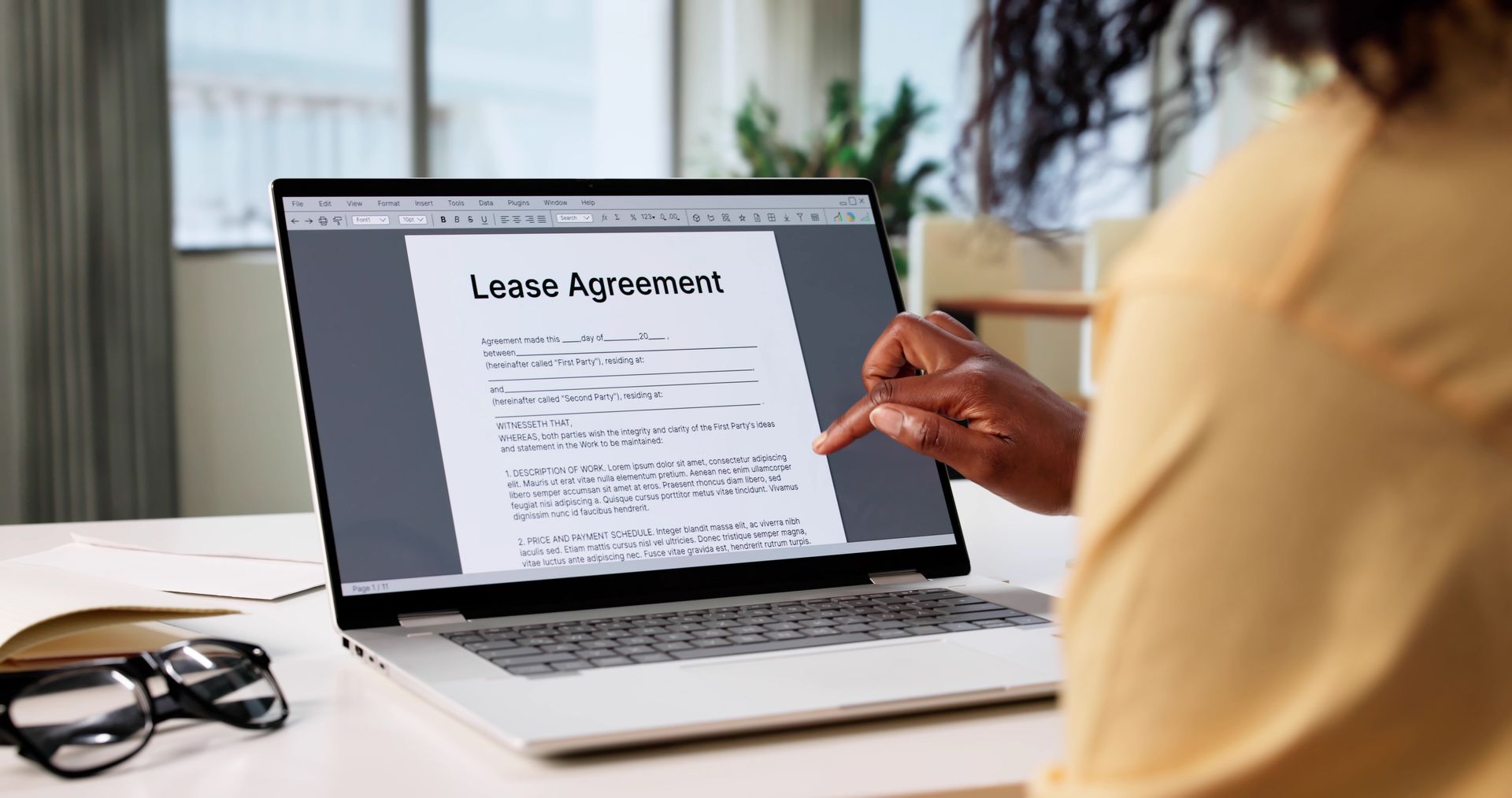 Person reviewing a lease agreement on a laptop at a desk, holding a pen in a bright office.