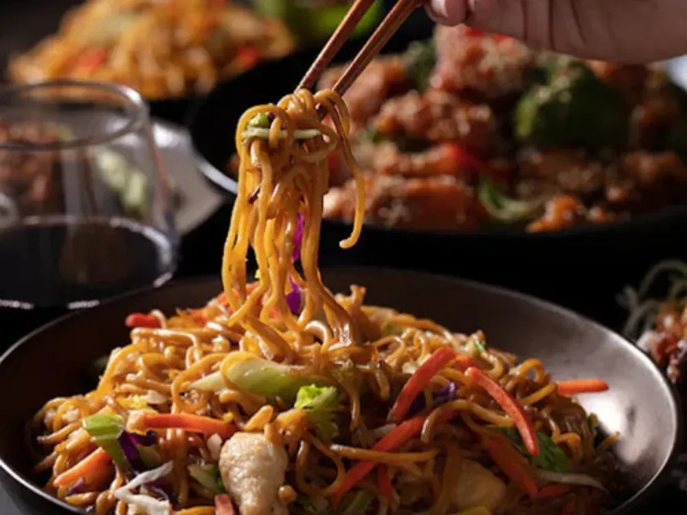 A person is holding chopsticks over a bowl of noodles and vegetables.