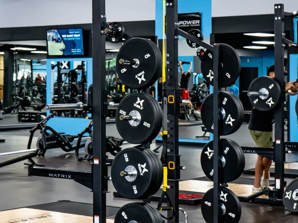 A man is squatting in a gym with a barbell rack filled with barbells.