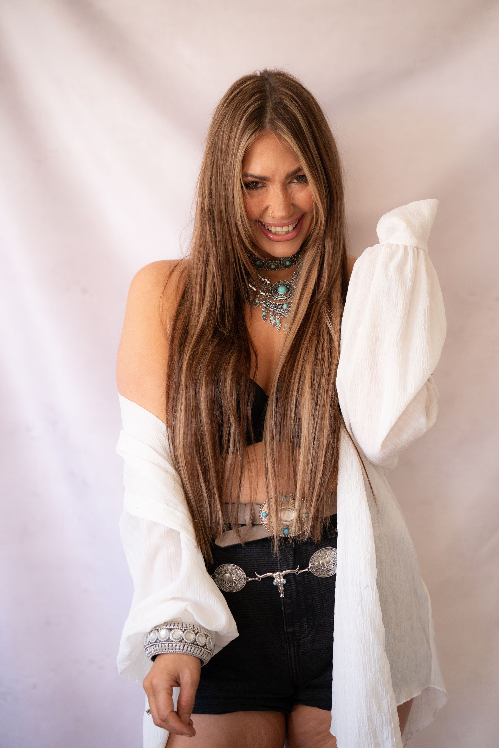 Woman with long hair smiles, wearing jewelry, bra, shorts, and white jacket.