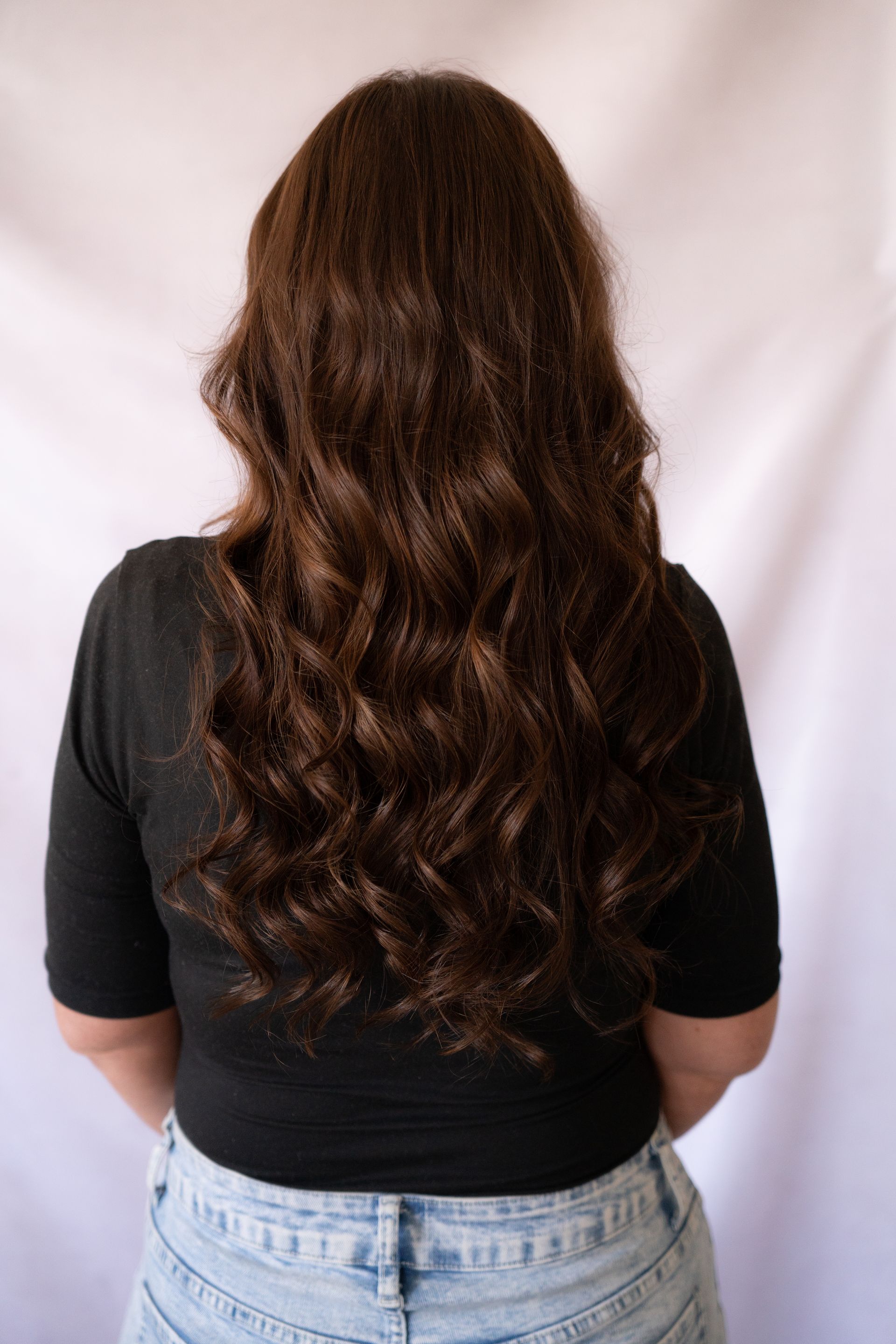 Woman with long, wavy brown hair, wearing a black shirt and jeans, standing against a white background.