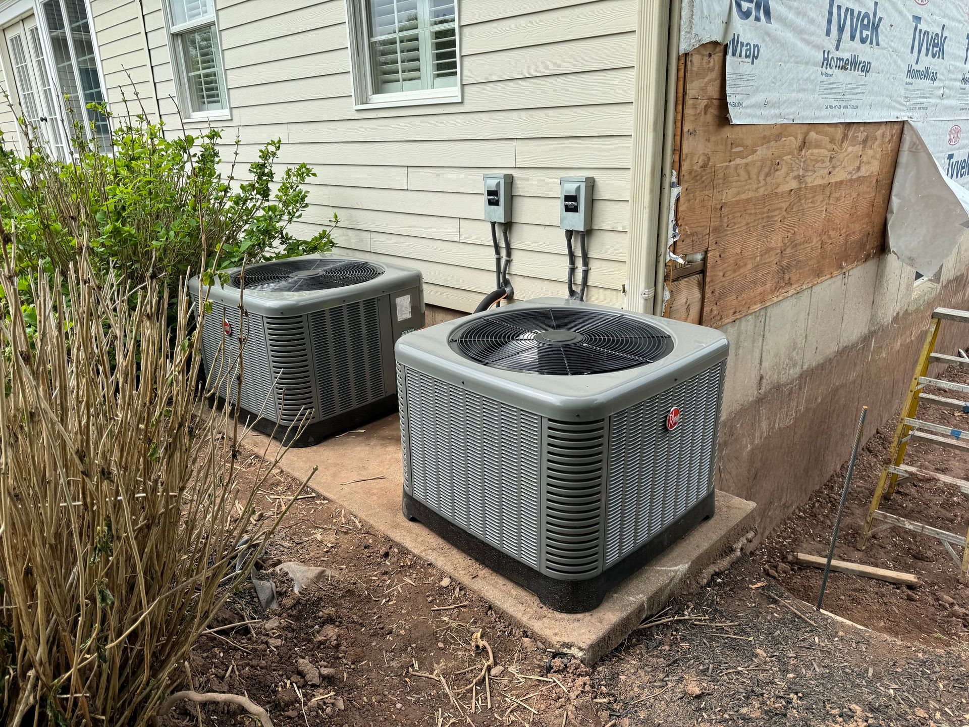 Two air conditioning units on concrete pads beside a building. Wiring boxes visible.