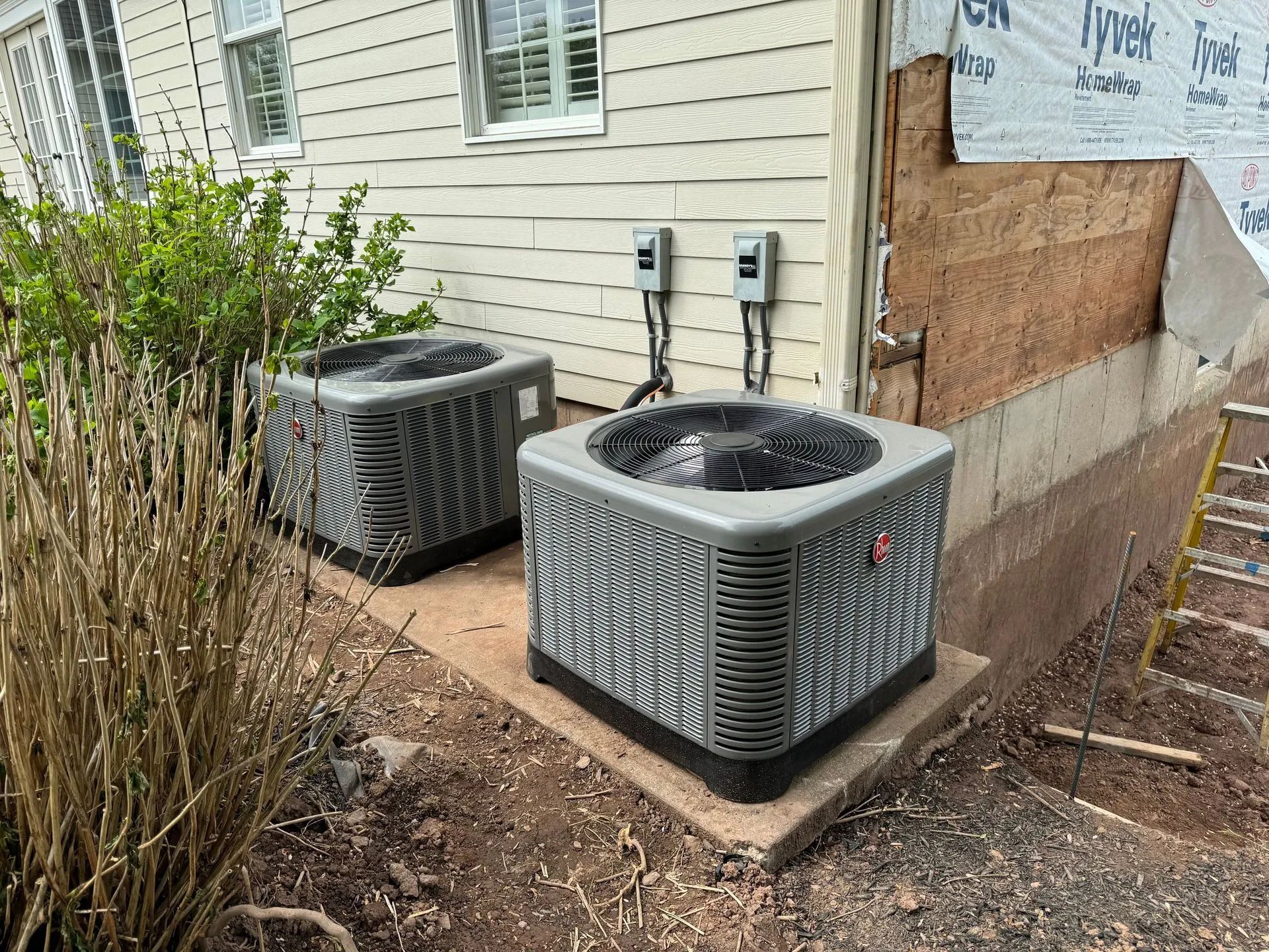 Two outdoor air conditioners beside a house, next to boarded-up siding and a concrete walkway.