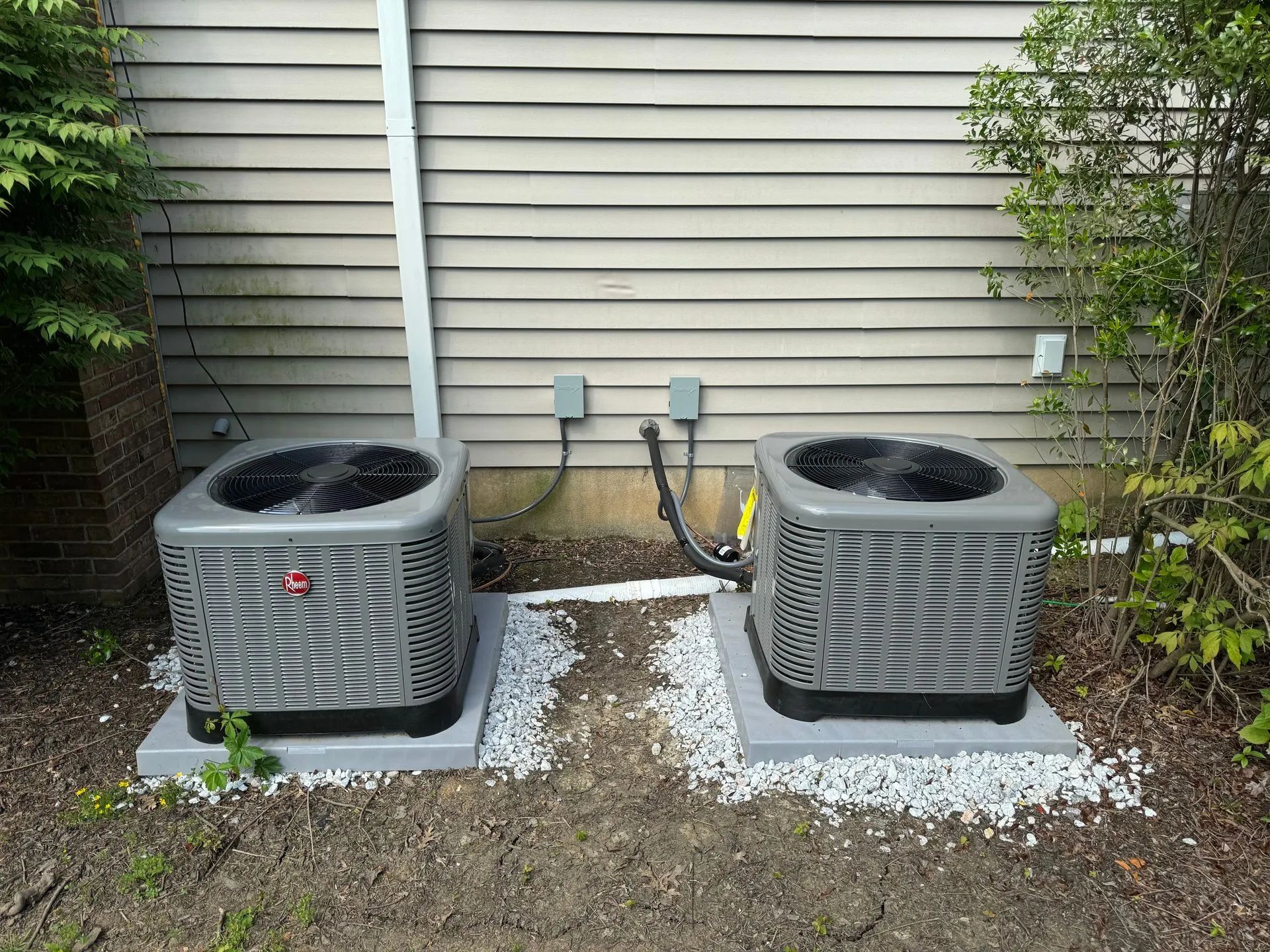 Two outdoor air conditioning units beside a house wall, surrounded by gravel and shrubs.