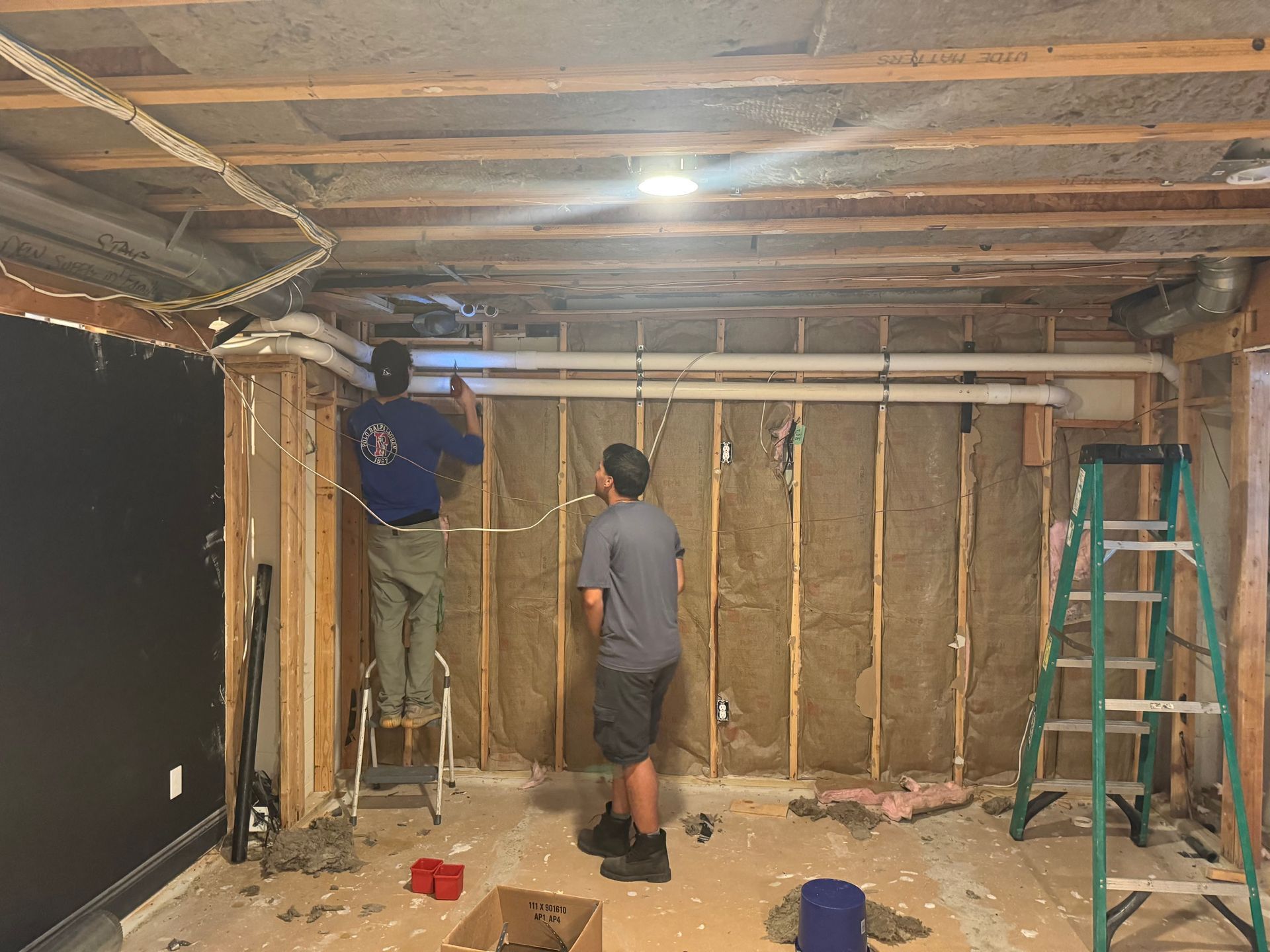 Two men installing PVC pipes and wiring on a partially constructed basement wall.