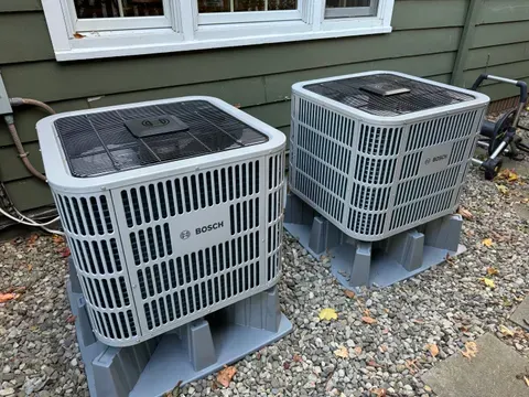 Two Bosch air conditioning units on gray platforms near a house.