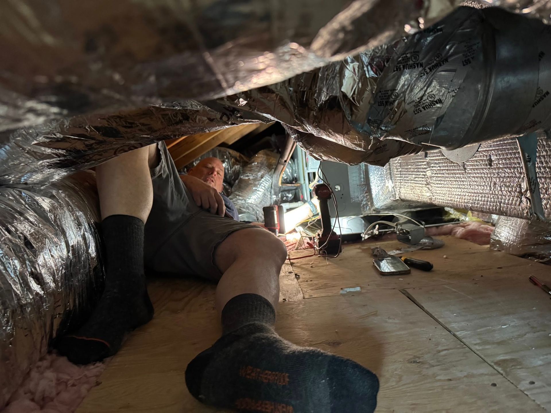 Person lying under ductwork, working on a mechanical system. Attentive, tool visible.