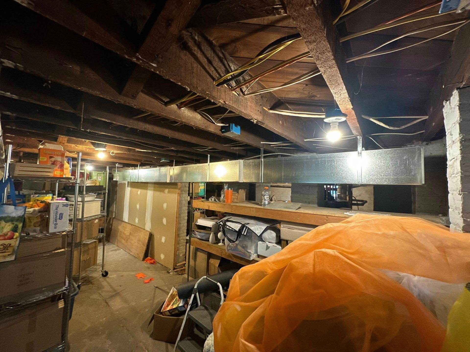 Basement with exposed beams, ductwork, storage shelves, and clutter, illuminated by overhead lights.