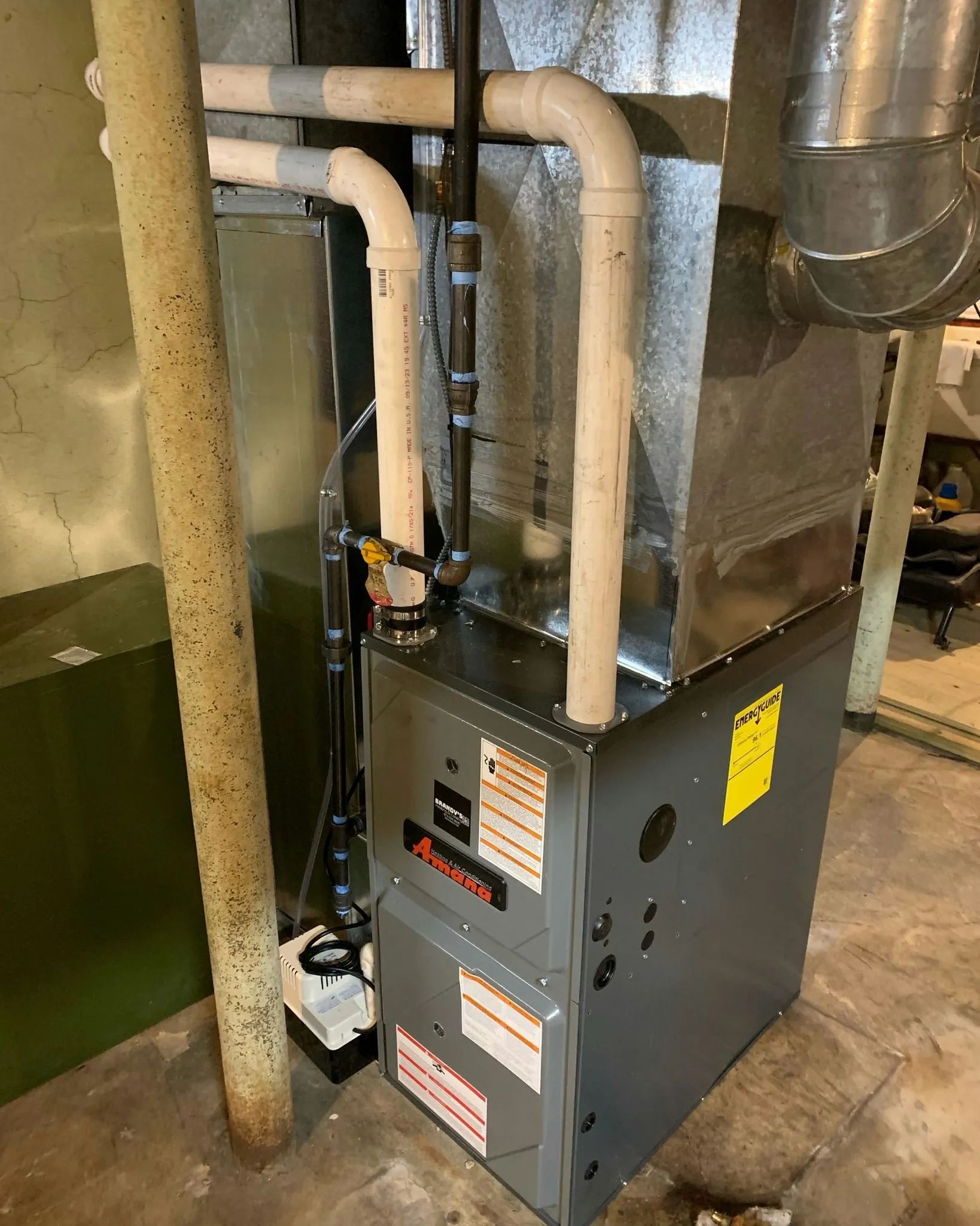Furnace in a basement with white PVC pipes, metal ductwork, and yellow energy efficiency sticker.