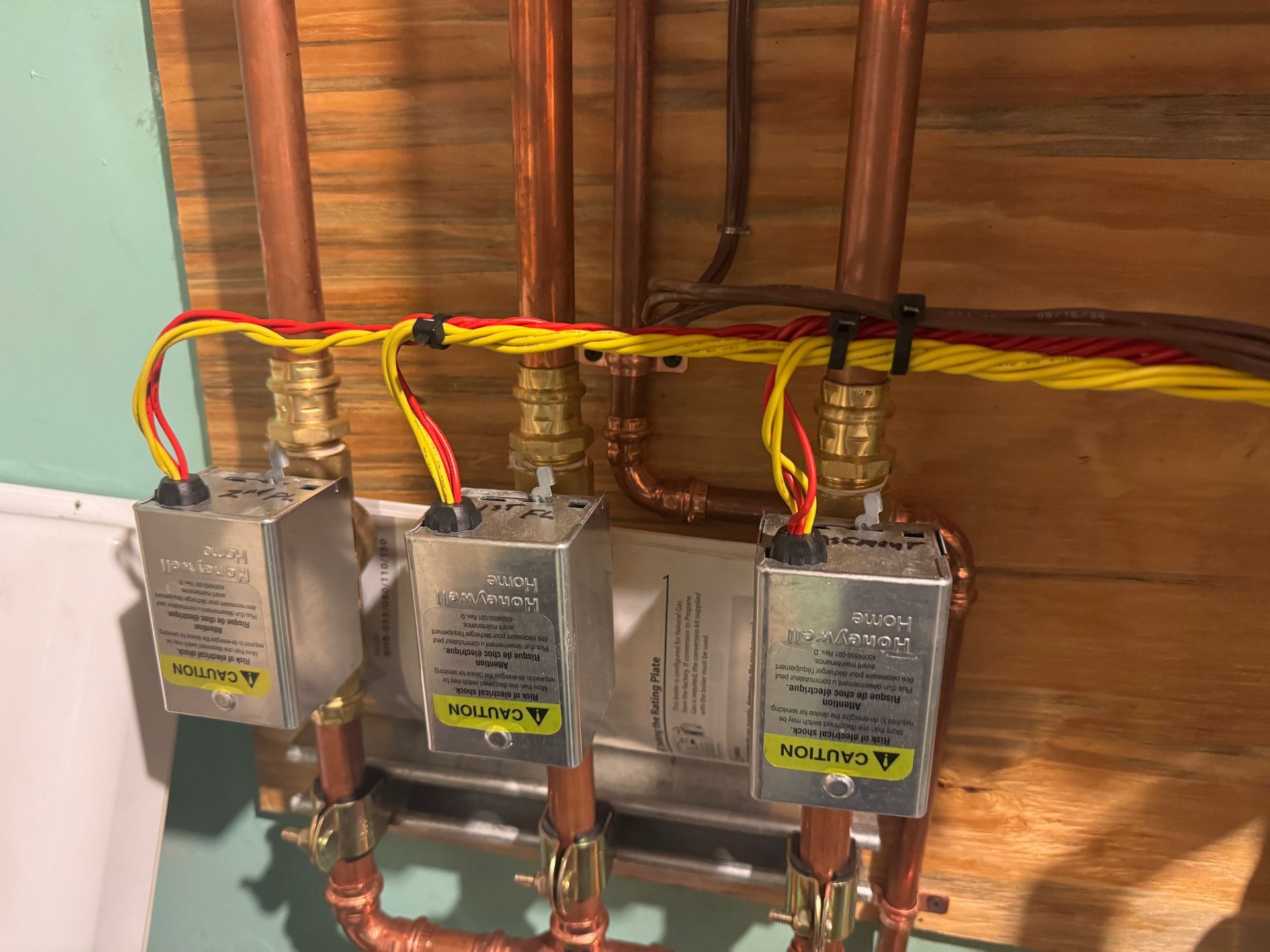 Copper pipes with three silver boxes and colored wires, secured with black zip ties, mounted on wood.