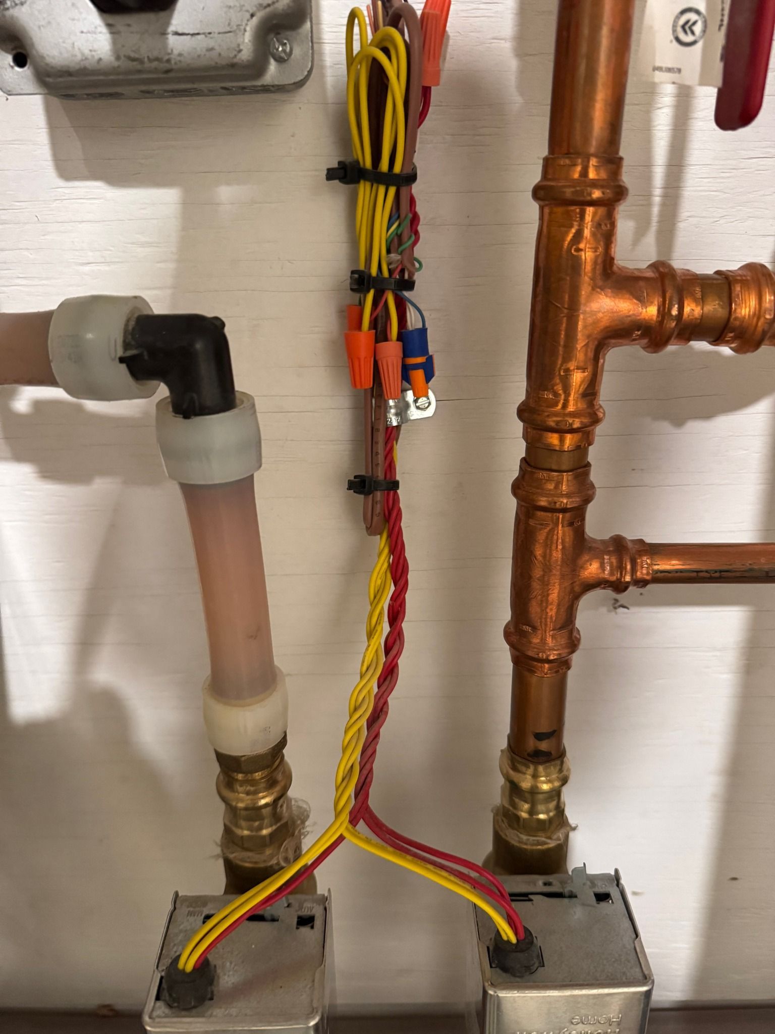 Copper pipes and electrical wiring connected to boxes on a white wall.
