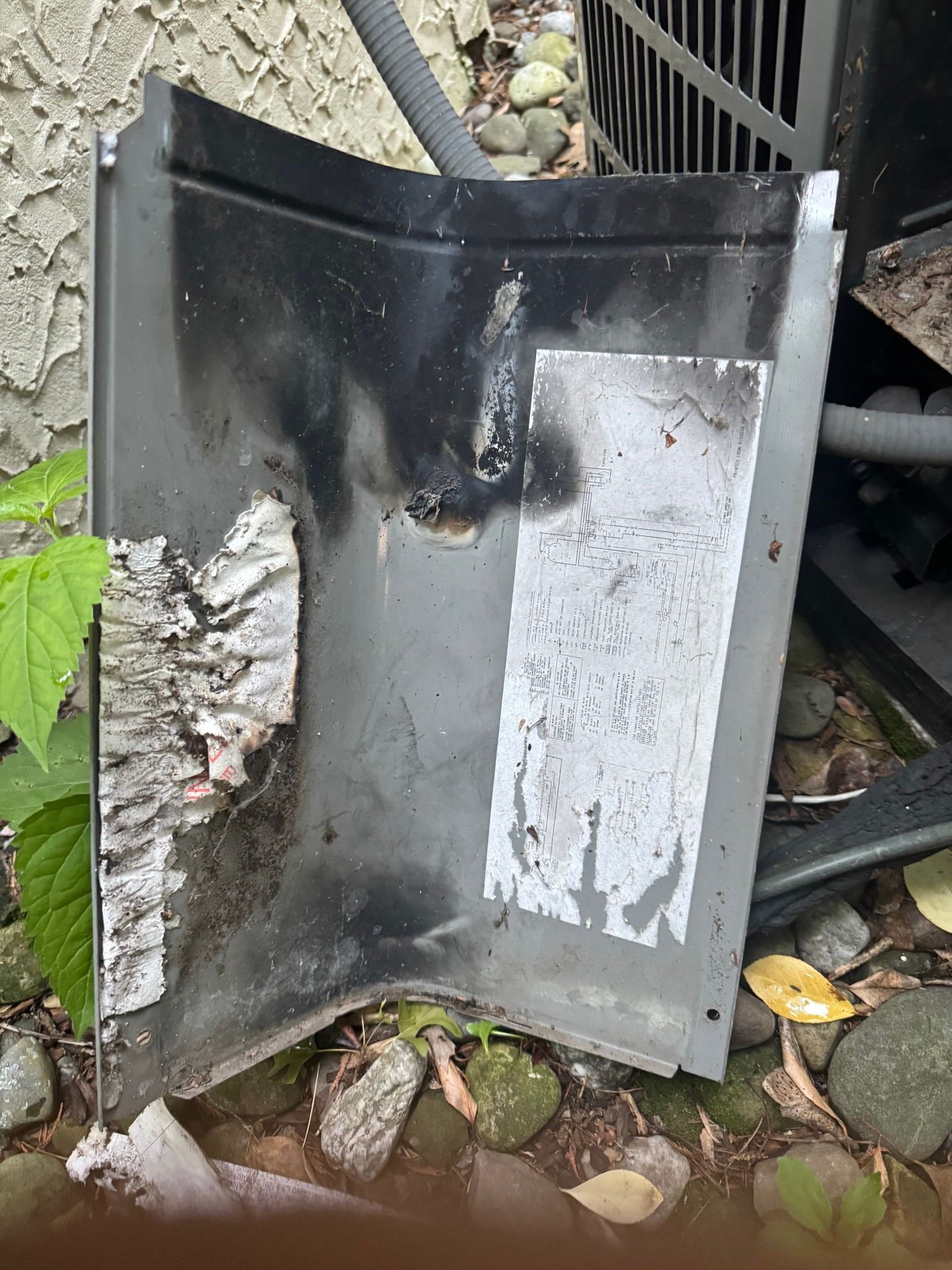 Damaged metal panel from an air conditioning unit. Black discoloration from likely fire damage. Outdoor setting with rocks.