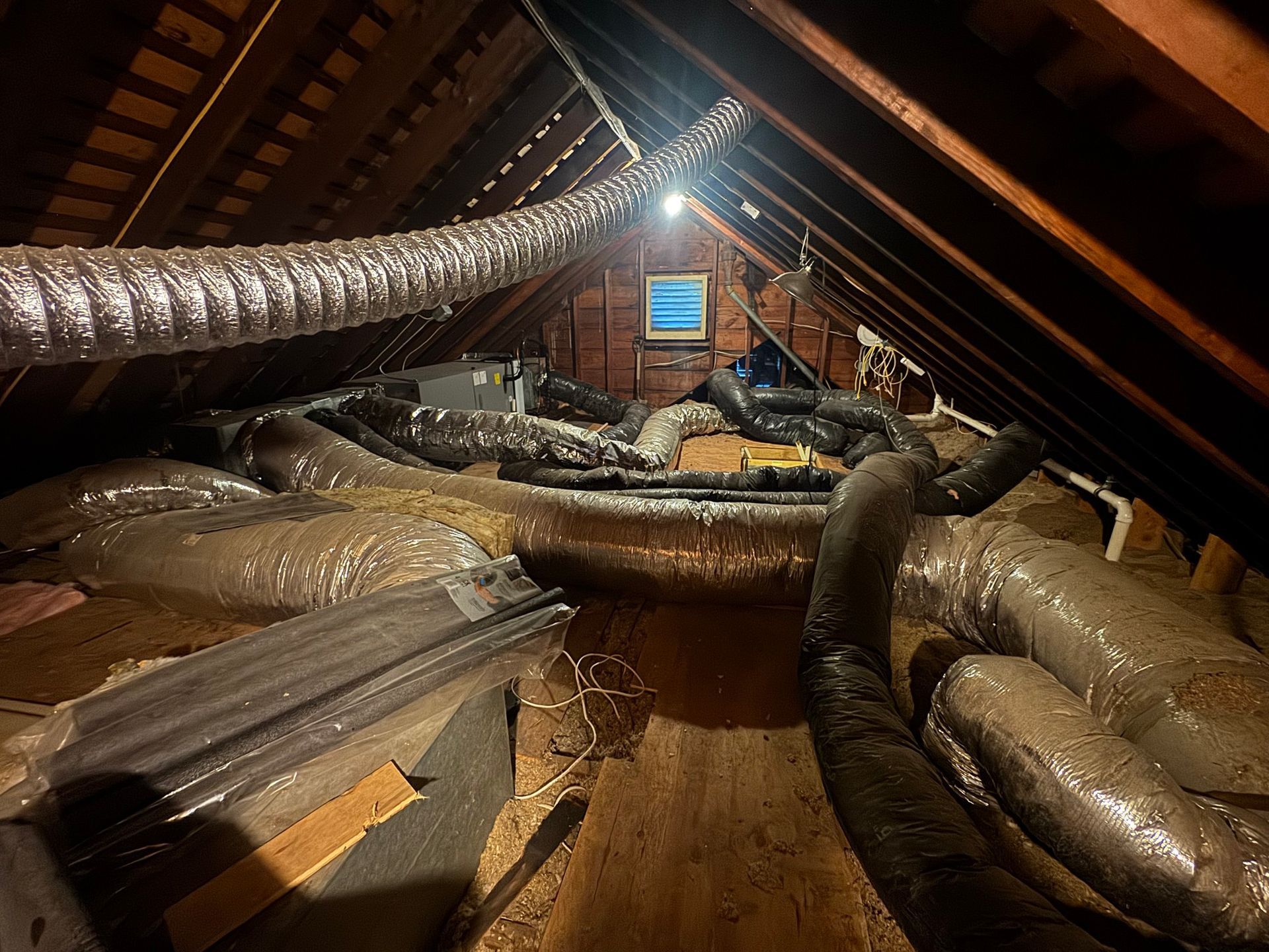 Attic with HVAC ductwork and a small window. Brown wooden beams, insulation, and flexible silver and black ducts.