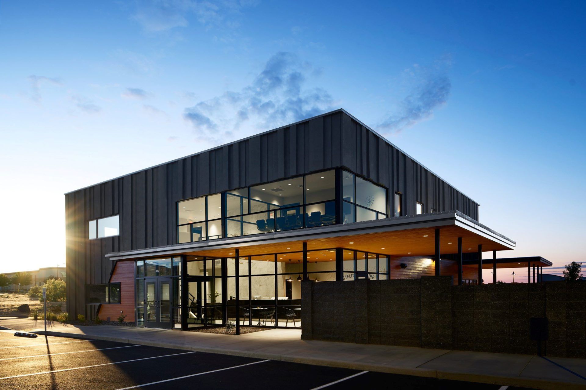 Modern building with glass front and flat roof, brick wall and parking lot, against a blue sky.