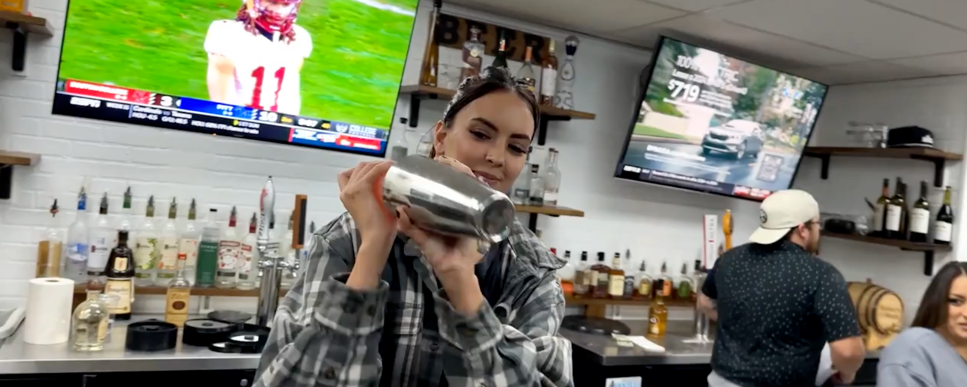 Bartender shaking a cocktail at a bar. Two TVs on the wall show sports. Other people are in the background.