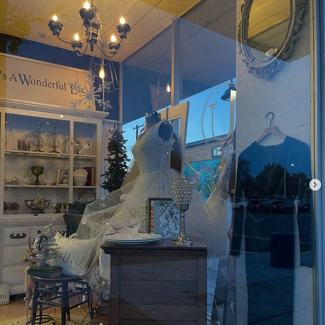 A bridal shop window display with a wedding dress on a mannequin, antique furniture, and a chandelier.
